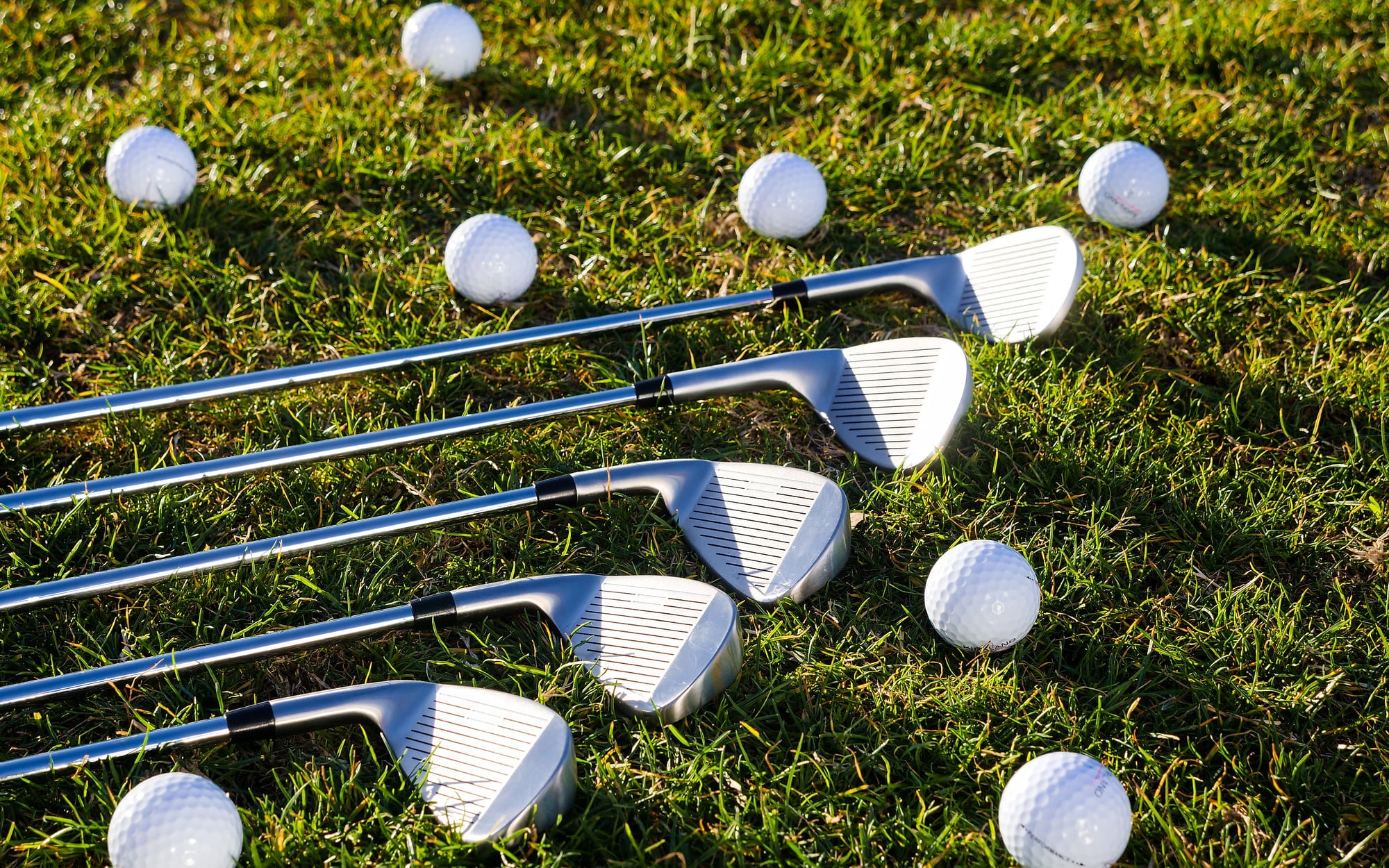Five iron heads laid out in a row on the turf with practice balls around them