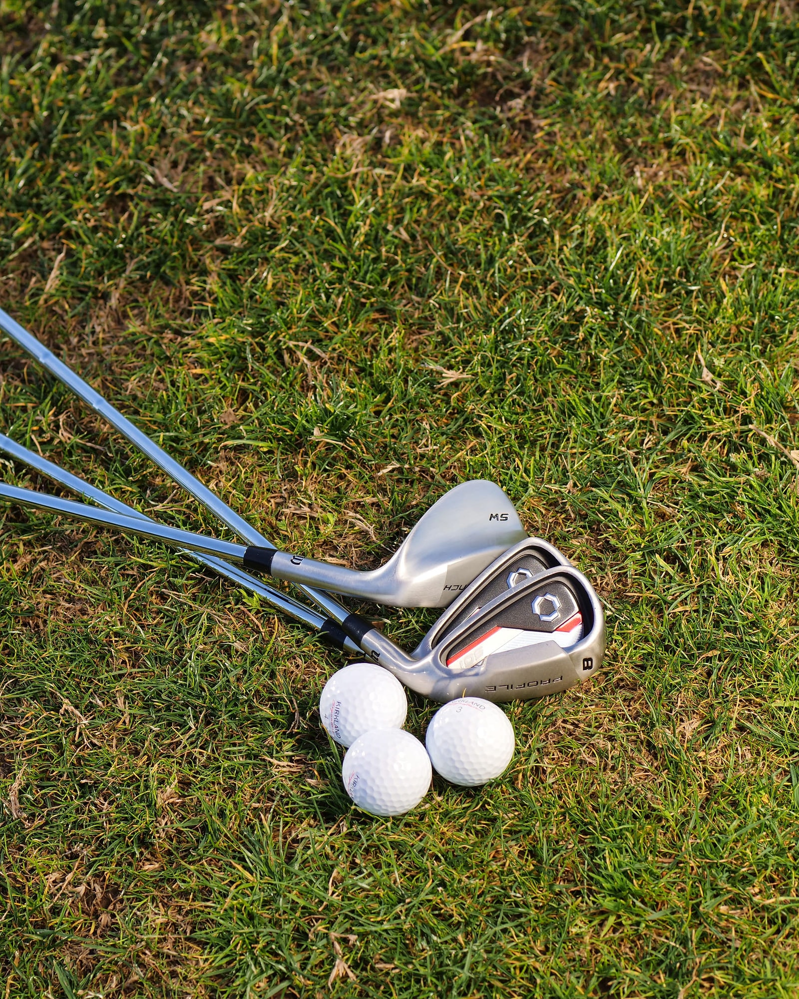 A set of irons laid out on cut grass beside three balls