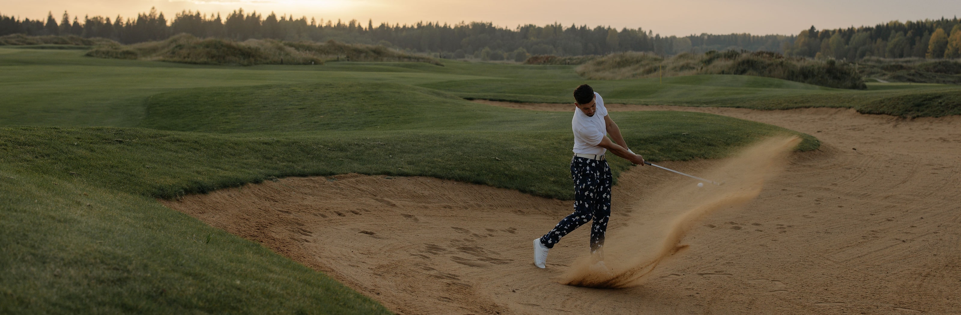 A golfer taking sand out of a bunker at dusk